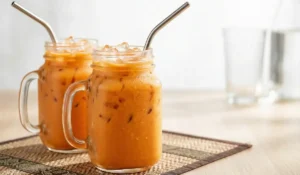 Zwei Henkelgläser gefüllt mit eiskaltem, authentischem Thai Eistee (Cha Yen) und Eiswürfeln, serviert mit Metallstrohhalmen auf einem geflochtenen Untersetzer.