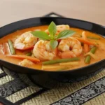 Gaeng Ped Gung ist ein authentisches rotes Thai Curry mit Garnelen, das reich an Aromen und Farben ist. Das Gericht enthält zarte Garnelen, lange grüne Bohnen, Bambussprossen und in Streifen geschnittene Karotten. Garniert mit frischem heiligem Basilikum, wird es in einem eleganten dunklen Teller präsentiert, der auf einer dekorativen Matte steht.