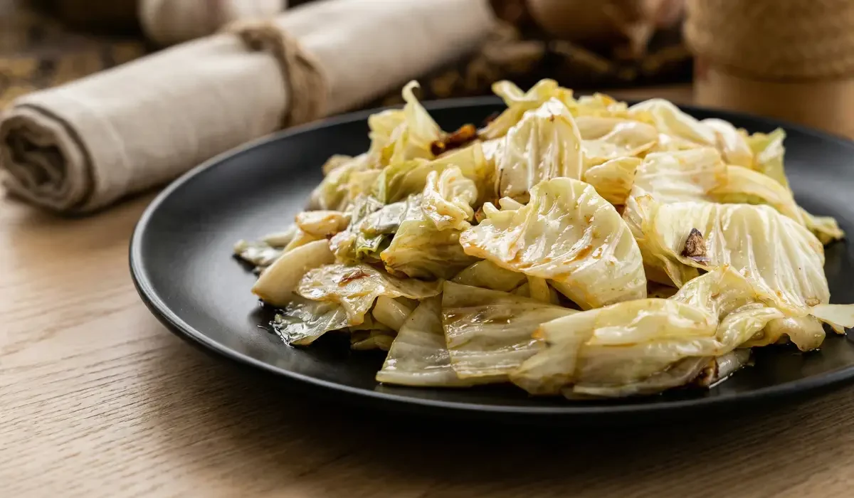 Nahaufnahme von einem schwarzen Teller auf einem Holztisch im hellen Tageslicht. Darauf liegt Gebratener Spitzkohl mit Fischsauce. Die mundgerechten Kohlstücke sind leicht glasig gebraten und weisen braune Röstaromen auf. Im unscharfen Hintergrund liegt eine rustikal gerollte Stoffserviette.