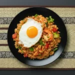 Ein Teller mit gebratenem Tom Yum Reis, der mit Garnelen, frischen Kräutern, Tomaten und einem Spiegelei garniert ist. Der Reis hat eine goldene Farbe und wird auf einem dekorativen Tischset präsentiert.