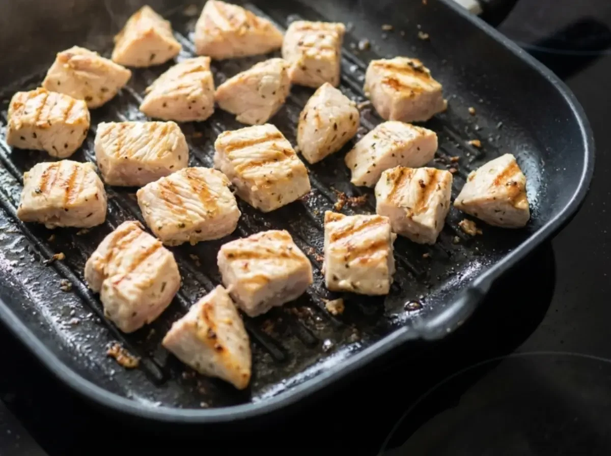 Angebratene Hähnchenwürfel mit Grillstreifen in einer Grillpfanne auf dem Herd.