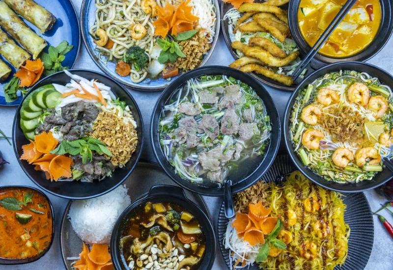 Eine vielfältige Auswahl an thailändischen Gerichten, die die Grundlagen der thailändischen Küche repräsentieren. Zu sehen sind verschiedene Schalen mit Reis, Nudeln, frischem Gemüse, Garnelen, Fleisch und aromatischen Kräutern, die zusammen ein farbenfrohes und einladendes Bild der thailändischen Gastronomie ergeben.