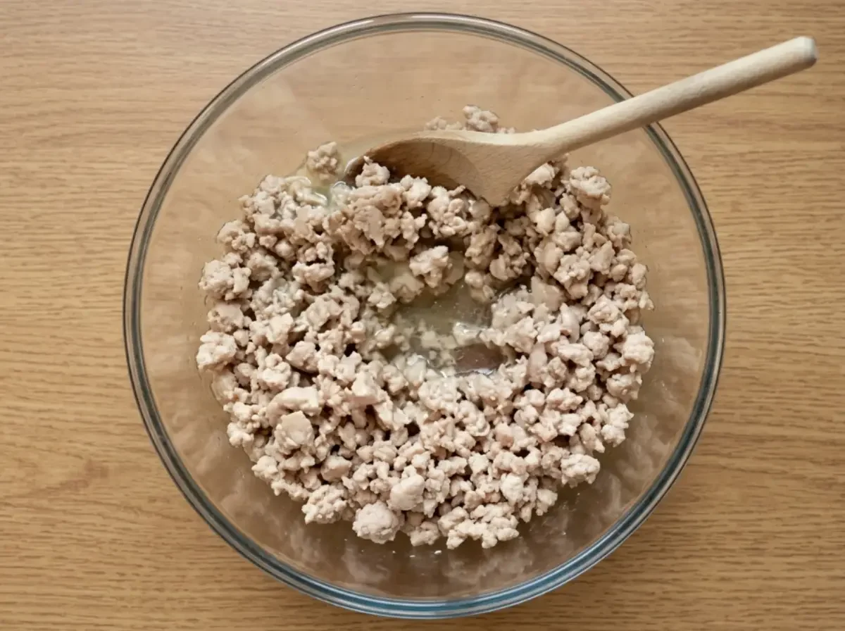 Gekochtes Schweinehackfleisch in seinem eigenen Saft in einer großen Glasschüssel mit einem Holzlöffel – der vorbereitete Fleisch-Grundstock für den Nam Sod.