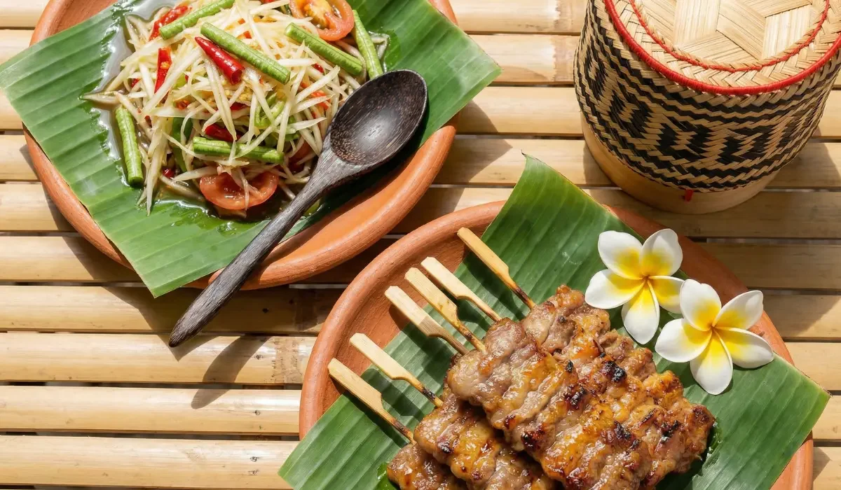 Moo Ping mit Papayasalat auf Bananenblättern angerichtet, daneben ein traditioneller Reisbehälter und tropische Blüten