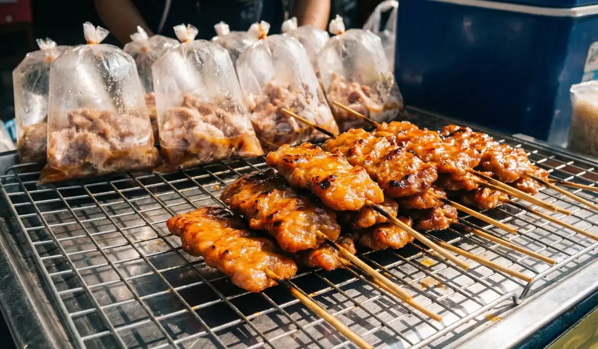 Frisch gegrillte Moo Ping Spieße auf einem Straßenmarkt, auf einem Metallrost mit mariniertem Fleisch im Hintergrund