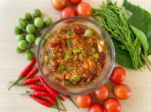 Nam Prik Kapi in Glasschale mit Chili und Gemüse, umgeben von Tomaten, Kräutern und grünen Auberginen.