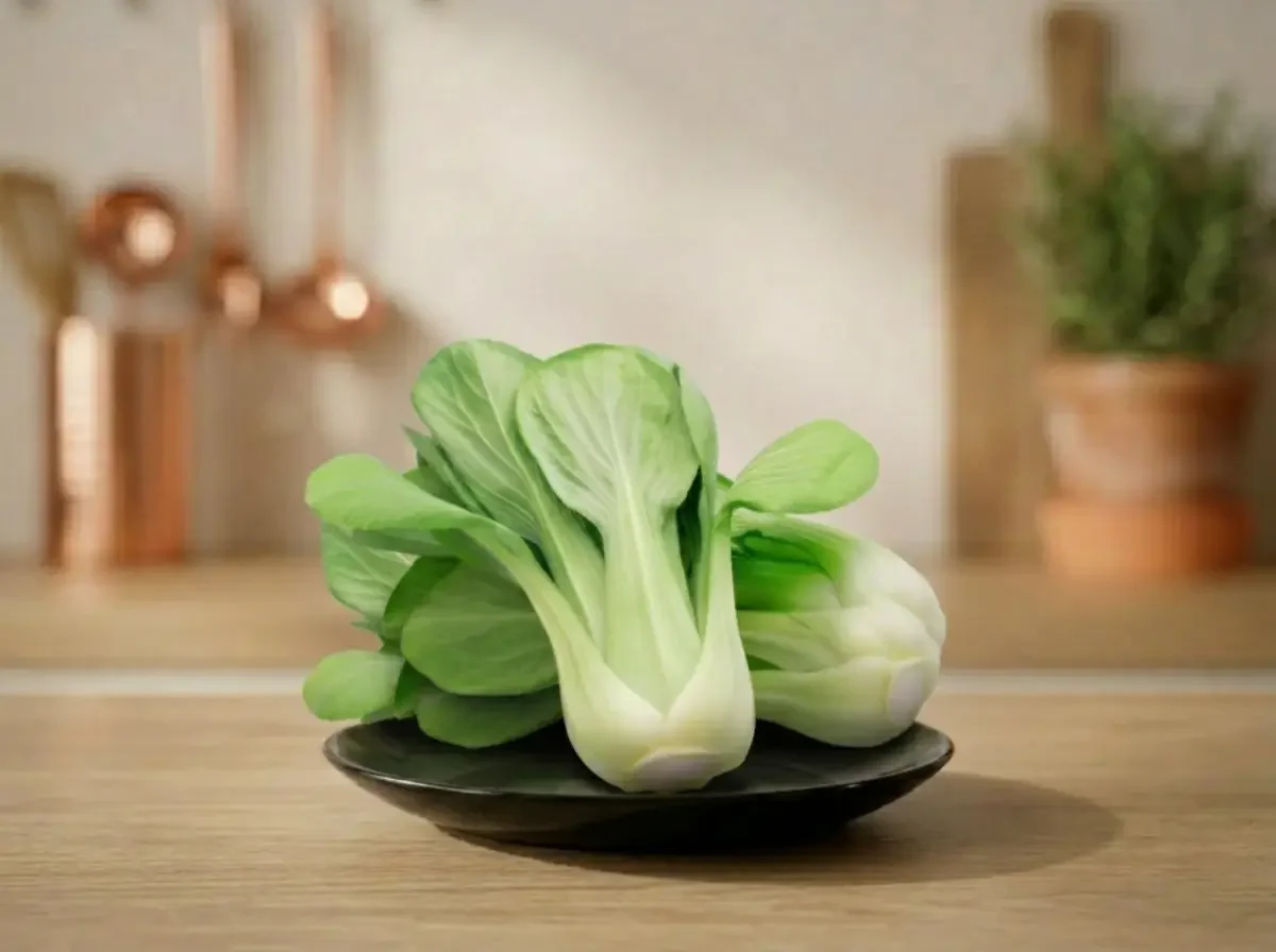 Zwei frische Köpfe Pak Choi liegen auf einem dunklen Teller auf einer hölzernen Arbeitsplatte in einer hellen Küche.