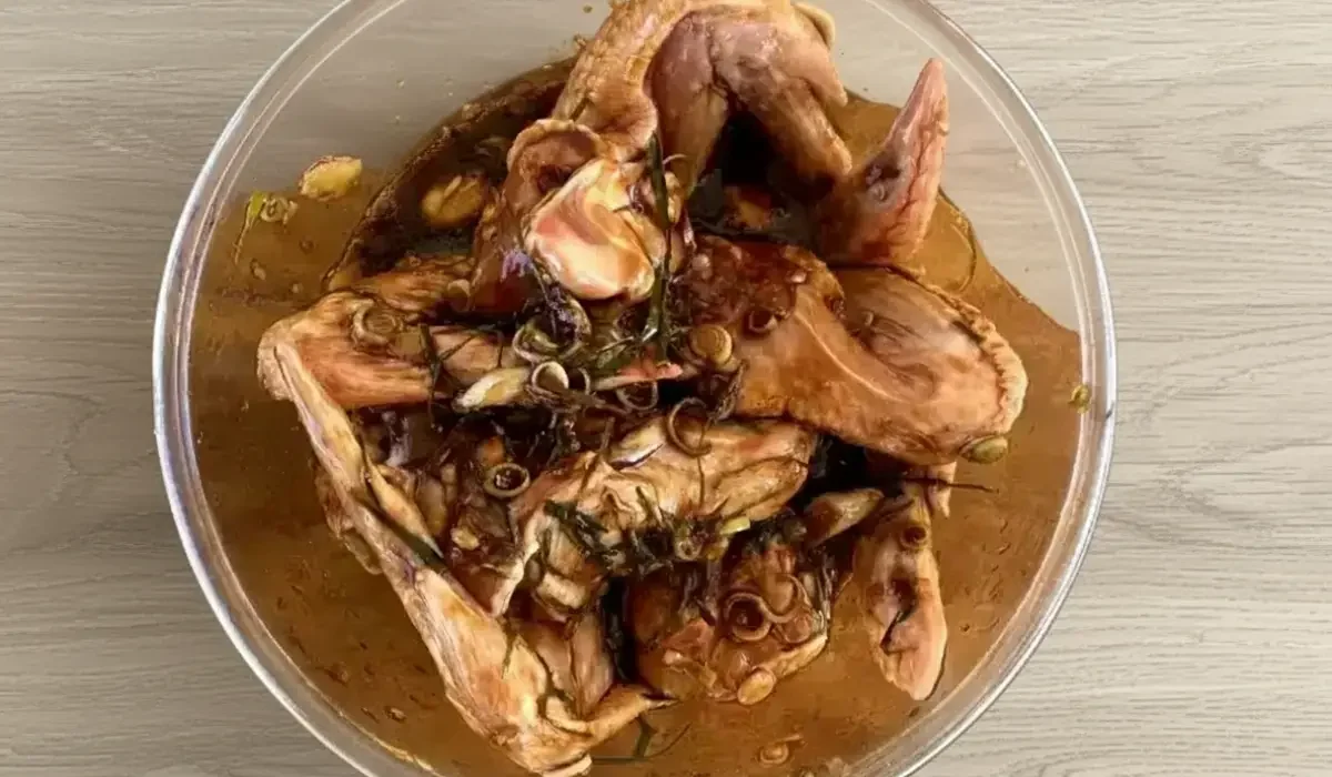 Rohe Thai Chicken Wings, die in einer Glasschüssel in einer dunklen, würzigen Marinade mit frischen Zitronengras-Ringen und Kaffir-Limettenblättern auf einem Tisch in Sonoma-Eiche-Optik eingelegt sind.