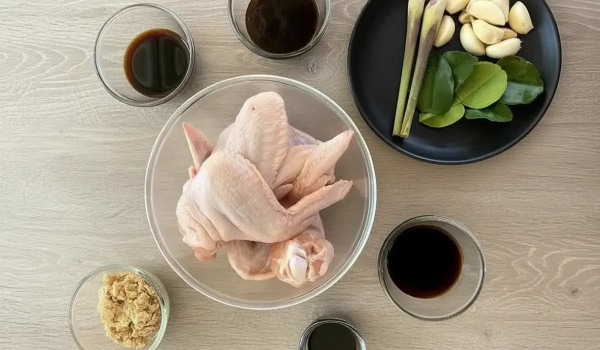 Mise-en-place der Zutaten für Thai Chicken Wings: Eine Schüssel mit rohem Hühnchen, umgeben von Schälchen mit Sojasaucen und Zucker sowie einem Teller mit frischem Zitronengras, Knoblauch und Limettenblättern.