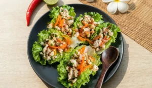 Vier thai Hähnchen Salat-Wraps (Larb Gai) auf einem mattschwarzen Teller. Die Salatblätter sind gefüllt mit Hähnchenhackfleisch, Karottenstreifen und Kräutern. Das Gericht steht auf einem hellen Holztisch, dekoriert mit einer roten Chilischote, einer Limette und weißen Frangipani-Blüten im Sonnenlicht.