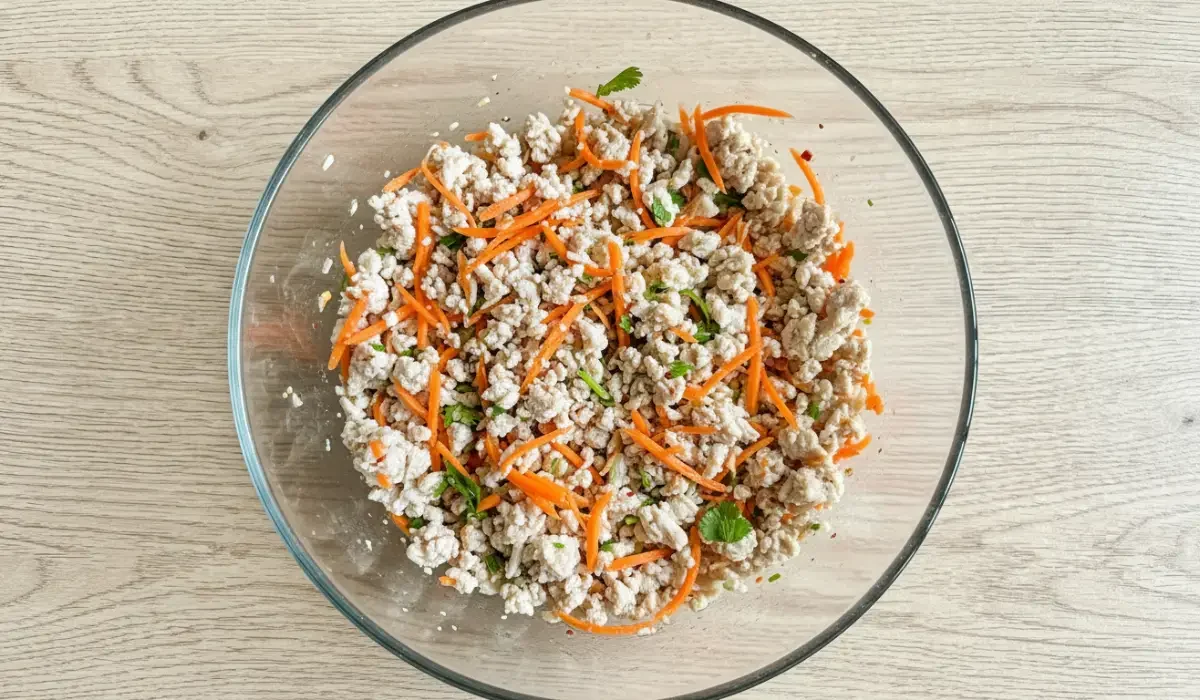 Eine Glasschüssel in der Draufsicht, gefüllt mit der Zubereitung für thailändischen Hähnchensalat. Man sieht eine Mischung aus krümeligem Hähnchenhackfleisch, frischen Karottenstiften und grünen Kräutern auf einem Tisch mit heller Holzoptik.