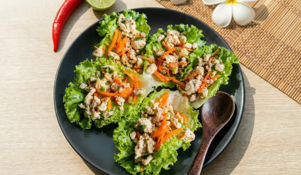 Vier thai Hähnchen Salat-Wraps (Larb Gai) auf einem mattschwarzen Teller. Die Salatblätter sind gefüllt mit Hähnchenhackfleisch, Karottenstreifen und Kräutern. Das Gericht steht auf einem hellen Holztisch, dekoriert mit einer roten Chilischote, einer Limette und weißen Frangipani-Blüten im Sonnenlicht.