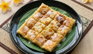 Thai Roti in Stücke geschnitten, goldbraun gebraten und mit süßer Milch beträufelt, serviert auf Bananenblatt auf schwarzem Teller.