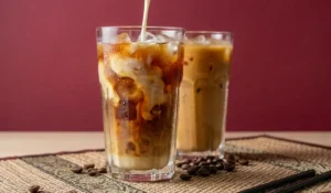 Thailändischer Eiskaffee in hohen Gläsern mit Eiswürfeln auf einem gewebten Tischset, wobei in das vordere Glas Milch eingegossen wird, die sich wirbelnd mit dem Kaffee vermischt; dekoriert mit Kaffeebohnen vor einem dunkelroten Hintergrund.