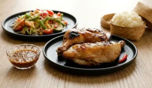 Thailändisches Grillhähnchen auf schwarzem Teller mit Dip-Sauce, Klebreis und Papayasalat