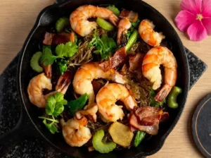 Blick von oben in eine gusseiserne Pfanne auf das thailändische Gericht Glasnudeln mit Garnelen. Die rosa Meeresfrüchte liegen appetitlich auf dunklen Nudeln, garniert mit Koriander, Sellerie und Speck.