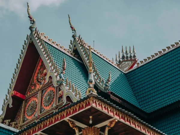 Kunstvoll verziertes thailändisches Tempeldach (Wat) mit grün-blauen Ziegeln, goldenen Giebel-Schnitzereien und thailändischen Naga-Verzierungen vor leicht bewölktem Himmel – symbolisch für thailändische Kultur und Geschichte.